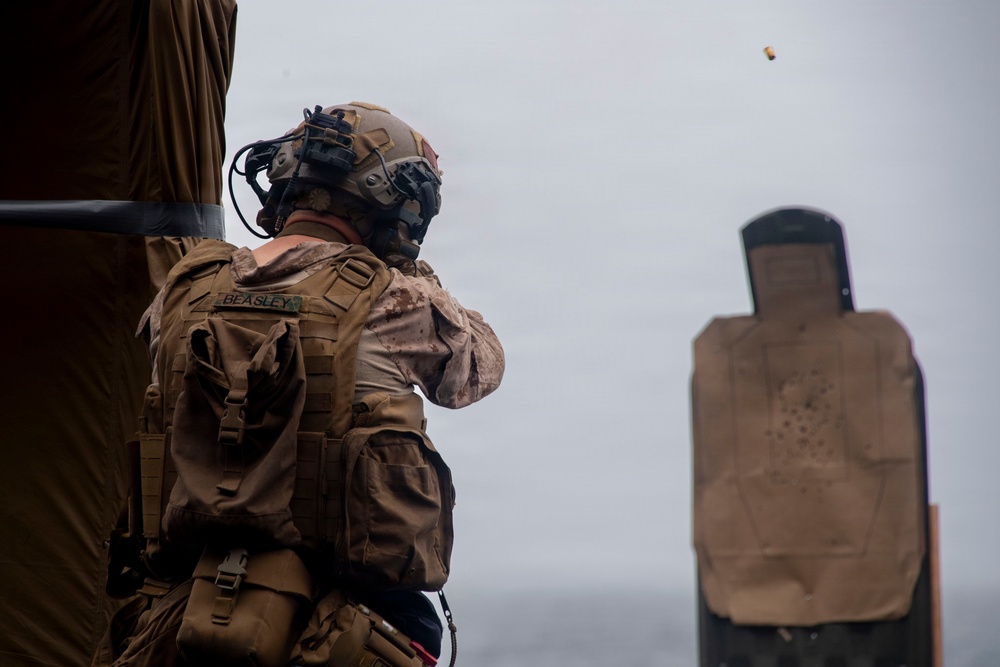 A military service member identified as 'Beasley' is engaged in a live-fire shooting exercise at a range. The shooter is wearing full tactical gear including a helmet with mounted equipment, plate carrier, and MARPAT camouflage uniform. A spent brass casing is visible in the air, and a paper silhouette target with visible bullet holes is downrange.