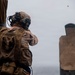 A military service member identified as 'Beasley' is engaged in a live-fire shooting exercise at a range. The shooter is wearing full tactical gear including a helmet with mounted equipment, plate carrier, and MARPAT camouflage uniform. A spent brass casing is visible in the air, and a paper silhouette target with visible bullet holes is downrange.