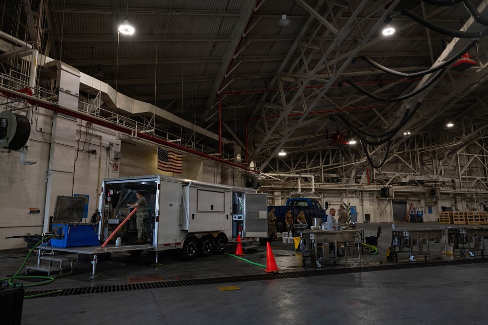 The image shows the interior of a large military hangar or maintenance facility with personnel conducting what appears to be equipment servicing or maintenance operations. A white enclosed trailer is positioned centrally, with several individuals in military uniforms and civilian attire working around it and nearby stainless steel work tables. An American flag is visible on the upper wall, confirming a U.S. military installation.