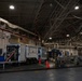 The image shows the interior of a large military hangar or maintenance facility with personnel conducting what appears to be equipment servicing or maintenance operations. A white enclosed trailer is positioned centrally, with several individuals in military uniforms and civilian attire working around it and nearby stainless steel work tables. An American flag is visible on the upper wall, confirming a U.S. military installation.