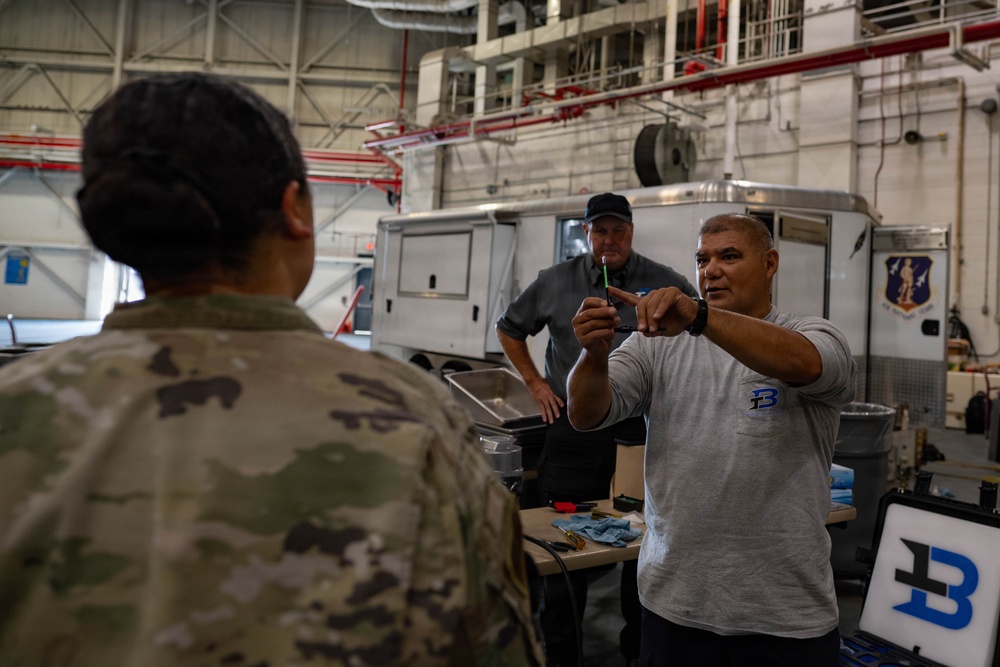 The image shows what appears to be a technical demonstration or training session inside a military hangar facility. A civilian contractor wearing a gray shirt with a 'B' logo is holding up a small component or tool while explaining something to a uniformed military personnel in camouflage (OCP/MultiCam pattern). A second civilian in a dark gray shirt and black cap observes from behind a work table.