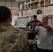 The image shows what appears to be a technical demonstration or training session inside a military hangar facility. A civilian contractor wearing a gray shirt with a 'B' logo is holding up a small component or tool while explaining something to a uniformed military personnel in camouflage (OCP/MultiCam pattern). A second civilian in a dark gray shirt and black cap observes from behind a work table.