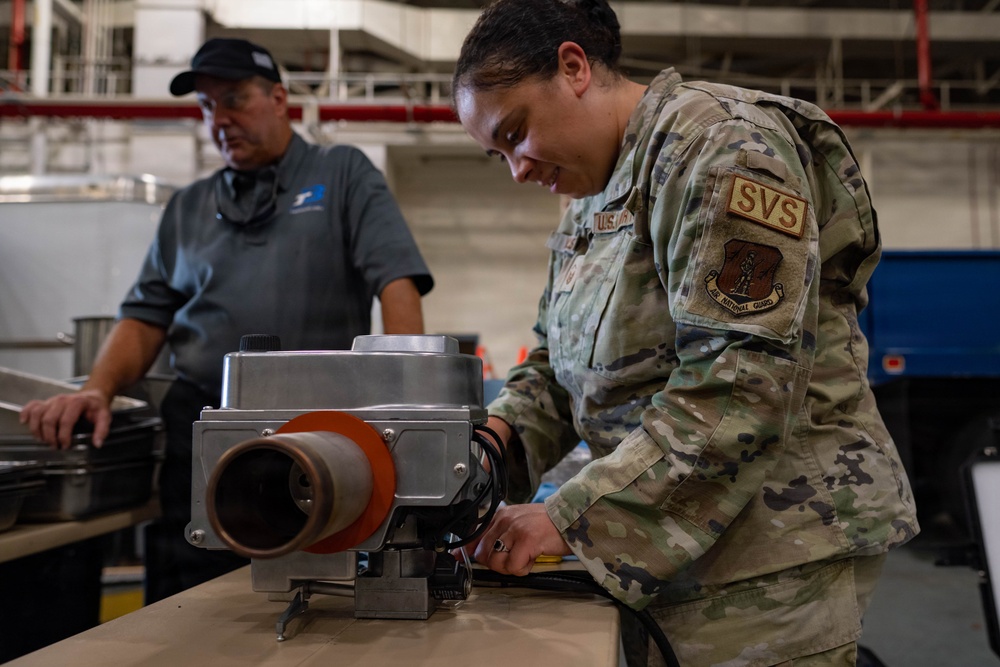 A U.S. Air National Guard service member wearing OCP (Operational Camouflage Pattern) uniform with 'SVS' patch is working with a piece of equipment on a workbench in what appears to be a hangar or industrial facility. A civilian contractor in a gray polo shirt and black cap observes or assists in the background. The scene appears to depict a maintenance or equipment setup operation.