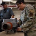 A U.S. Air National Guard service member wearing OCP (Operational Camouflage Pattern) uniform with 'SVS' patch is working with a piece of equipment on a workbench in what appears to be a hangar or industrial facility. A civilian contractor in a gray polo shirt and black cap observes or assists in the background. The scene appears to depict a maintenance or equipment setup operation.