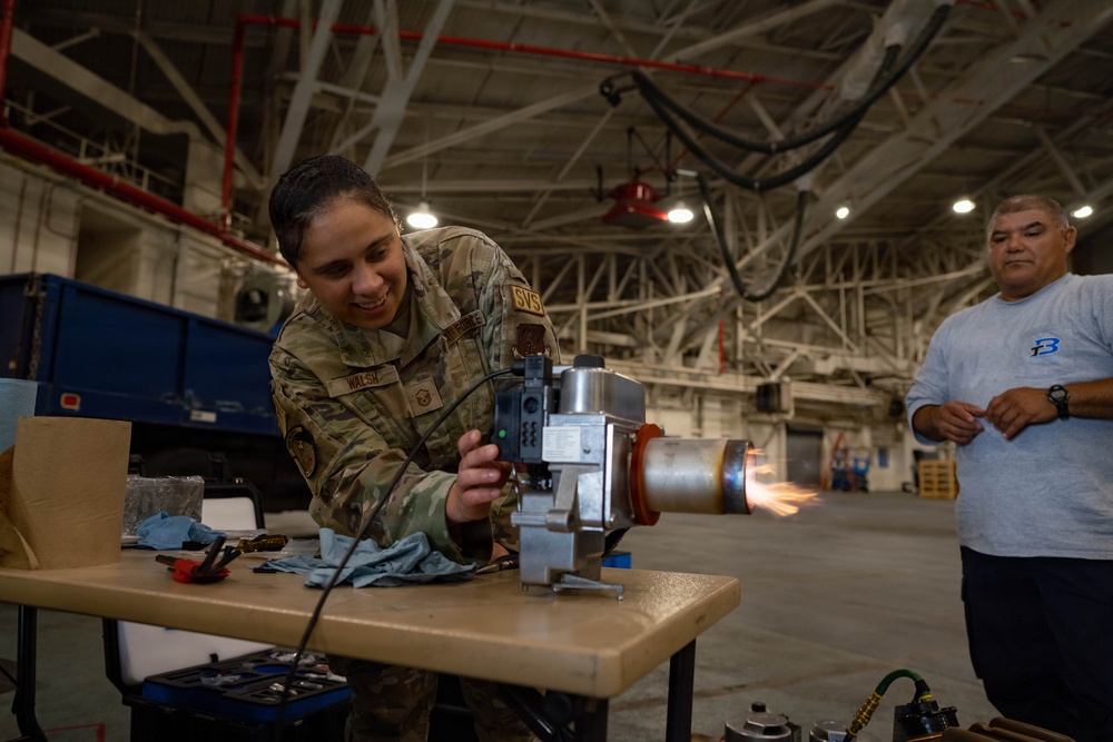 A U.S. Air Force service member wearing OCP (Operational Camouflage Pattern) uniform with name tape reading 'WALSH' and 'SVS' patch is working on a piece of mechanical equipment at a workbench inside a large military hangar. A civilian male in a gray shirt observes from the background. Sparks or a flame are visible in the mid-background, suggesting additional equipment testing is occurring simultaneously.