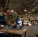 A U.S. Air Force service member wearing OCP (Operational Camouflage Pattern) uniform with name tape reading 'WALSH' and 'SVS' patch is working on a piece of mechanical equipment at a workbench inside a large military hangar. A civilian male in a gray shirt observes from the background. Sparks or a flame are visible in the mid-background, suggesting additional equipment testing is occurring simultaneously.