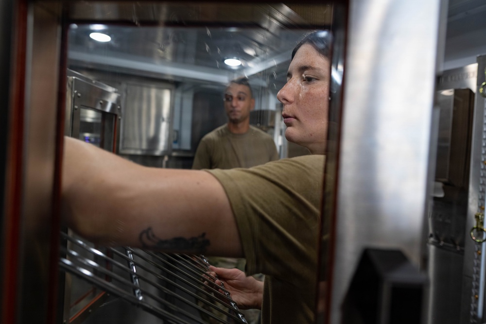 A woman in olive/military-style clothing reaches into what appears to be a commercial oven or warming unit, adjusting metal racks inside. A man in similar olive attire stands in the background observing. The setting appears to be a military or institutional kitchen facility.