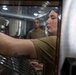 A woman in olive/military-style clothing reaches into what appears to be a commercial oven or warming unit, adjusting metal racks inside. A man in similar olive attire stands in the background observing. The setting appears to be a military or institutional kitchen facility.