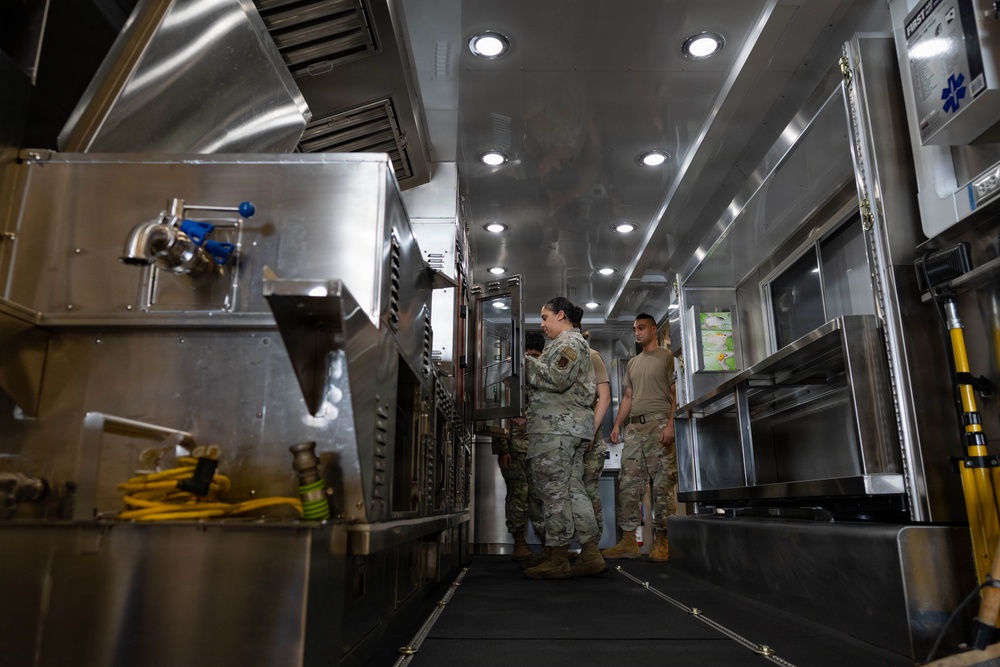 Two military personnel in camouflage uniforms are inspecting or operating equipment inside what appears to be a mobile military field kitchen unit. The interior is fitted with stainless steel commercial-grade cooking equipment, ventilation hoods, and recessed lighting. A first aid kit sign is visible on the right side wall, indicating this is a functional operational support vehicle.