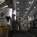 Two military personnel in camouflage uniforms are inspecting or operating equipment inside what appears to be a mobile military field kitchen unit. The interior is fitted with stainless steel commercial-grade cooking equipment, ventilation hoods, and recessed lighting. A first aid kit sign is visible on the right side wall, indicating this is a functional operational support vehicle.