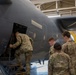 Military personnel in camouflage uniforms are inspecting or accessing a large gray military aircraft inside a hangar. One service member is leaning into an open access door or hatch on the fuselage while three others observe. The setting appears to be a maintenance or familiarization activity conducted in an indoor aircraft hangar.