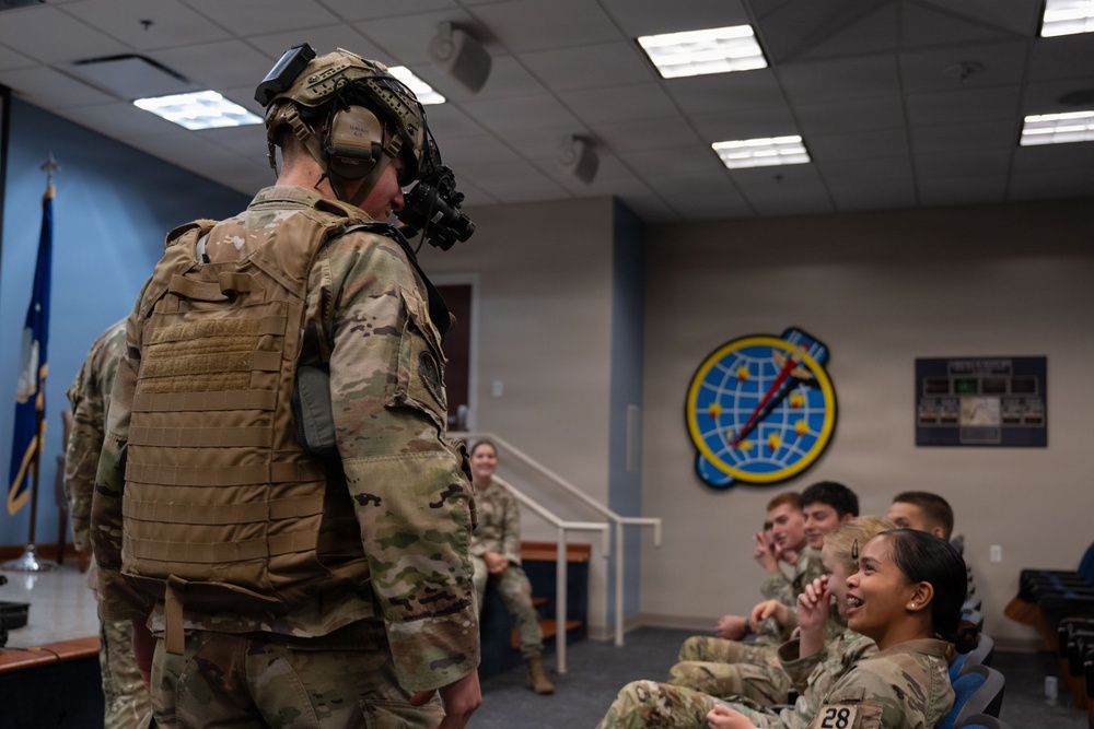 A fully equipped military personnel wearing tactical gear, helmet with night vision device, plate carrier vest, and communications headset stands before a group of seated military trainees or students in a briefing room. The audience appears engaged and amused, with several individuals smiling and reacting positively. A unit emblem featuring a globe and lightning bolt is visible on the wall in the background.