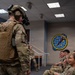 A fully equipped military personnel wearing tactical gear, helmet with night vision device, plate carrier vest, and communications headset stands before a group of seated military trainees or students in a briefing room. The audience appears engaged and amused, with several individuals smiling and reacting positively. A unit emblem featuring a globe and lightning bolt is visible on the wall in the background.