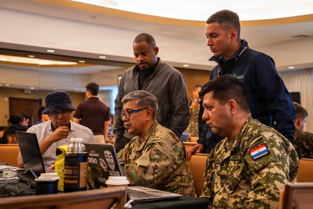 A group of five individuals, including two military personnel in camouflage uniforms and three civilians, are gathered around laptops in what appears to be a conference or training room. One soldier displays a Paraguay flag patch on their uniform, suggesting an international military or cybersecurity exercise. The group appears to be collaborating on a technical task displayed on the laptop screens.
