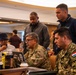A group of five individuals, including two military personnel in camouflage uniforms and three civilians, are gathered around laptops in what appears to be a conference or training room. One soldier displays a Paraguay flag patch on their uniform, suggesting an international military or cybersecurity exercise. The group appears to be collaborating on a technical task displayed on the laptop screens.