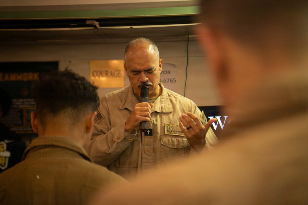 A military chaplain in tan/desert camouflage uniform is speaking or praying into a microphone, with a cross insignia visible on his uniform. He appears to be leading a religious service or prayer gathering, with other uniformed personnel visible in the foreground with their backs to the camera. The setting appears to be an indoor military facility with motivational signage visible in the background.