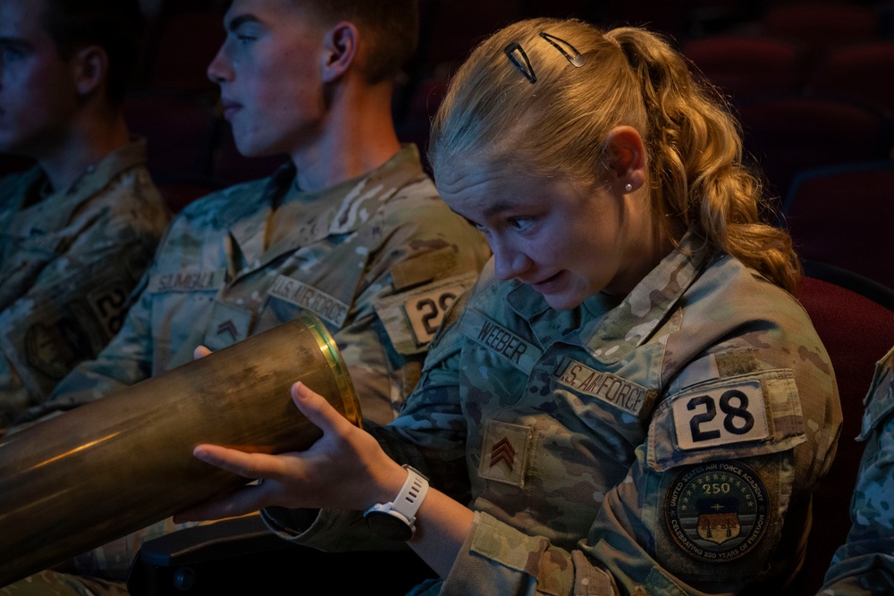 A U.S. Air Force Academy cadet named Weeber, wearing squadron number 28, examines a large brass cylindrical object while seated in what appears to be an auditorium or theater. She is wearing OCP (Operational Camouflage Pattern) uniform with a Corporal rank insignia and a United States Air Force Academy patch celebrating 250 years. Other cadets in similar uniforms are visible seated beside her.