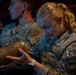 A U.S. Air Force Academy cadet named Weeber, wearing squadron number 28, examines a large brass cylindrical object while seated in what appears to be an auditorium or theater. She is wearing OCP (Operational Camouflage Pattern) uniform with a Corporal rank insignia and a United States Air Force Academy patch celebrating 250 years. Other cadets in similar uniforms are visible seated beside her.