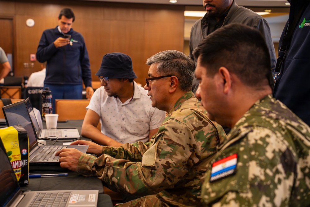 A group of individuals, including at least two military personnel in camouflage uniforms, are gathered around laptops in what appears to be a conference or training room. One soldier wearing glasses is actively typing on a laptop while others observe. The setting suggests a cybersecurity exercise or military technology training event.