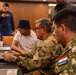 A group of individuals, including at least two military personnel in camouflage uniforms, are gathered around laptops in what appears to be a conference or training room. One soldier wearing glasses is actively typing on a laptop while others observe. The setting suggests a cybersecurity exercise or military technology training event.
