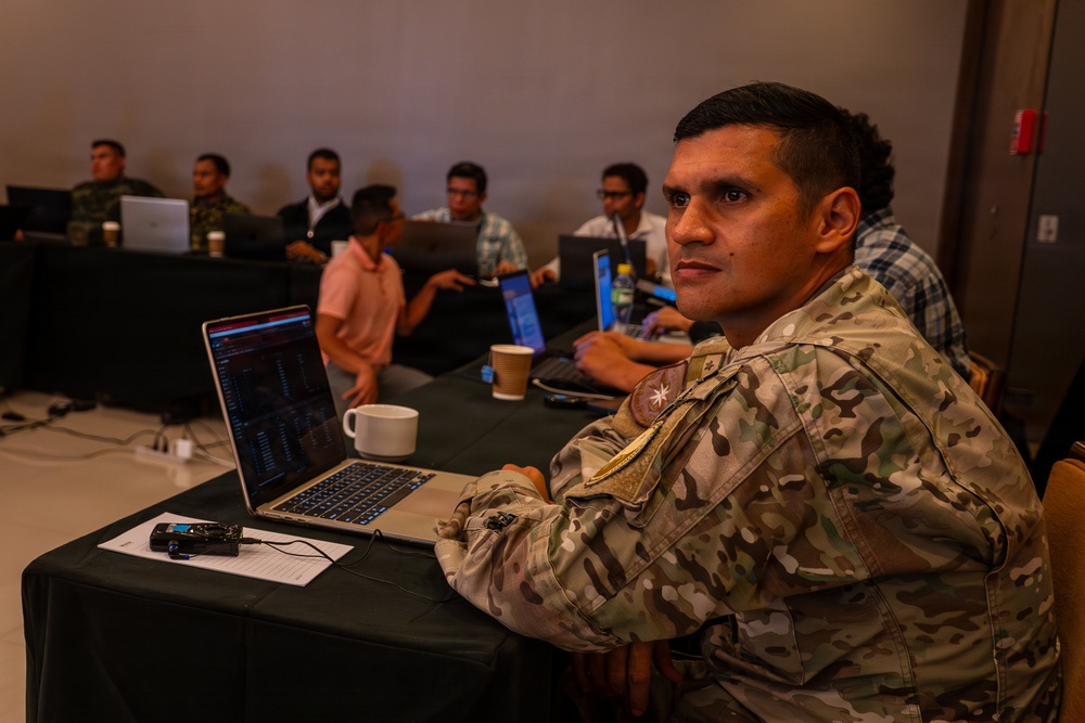 A military personnel member in camouflage uniform sits at a table with a laptop and coffee cup, looking toward the camera during what appears to be a training or conference session. Several other individuals, some in military uniforms and some in civilian clothing, are seated at tables in the background working on laptops. The setting appears to be a conference room or training facility with dim ambient lighting.