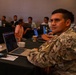 A military personnel member in camouflage uniform sits at a table with a laptop and coffee cup, looking toward the camera during what appears to be a training or conference session. Several other individuals, some in military uniforms and some in civilian clothing, are seated at tables in the background working on laptops. The setting appears to be a conference room or training facility with dim ambient lighting.