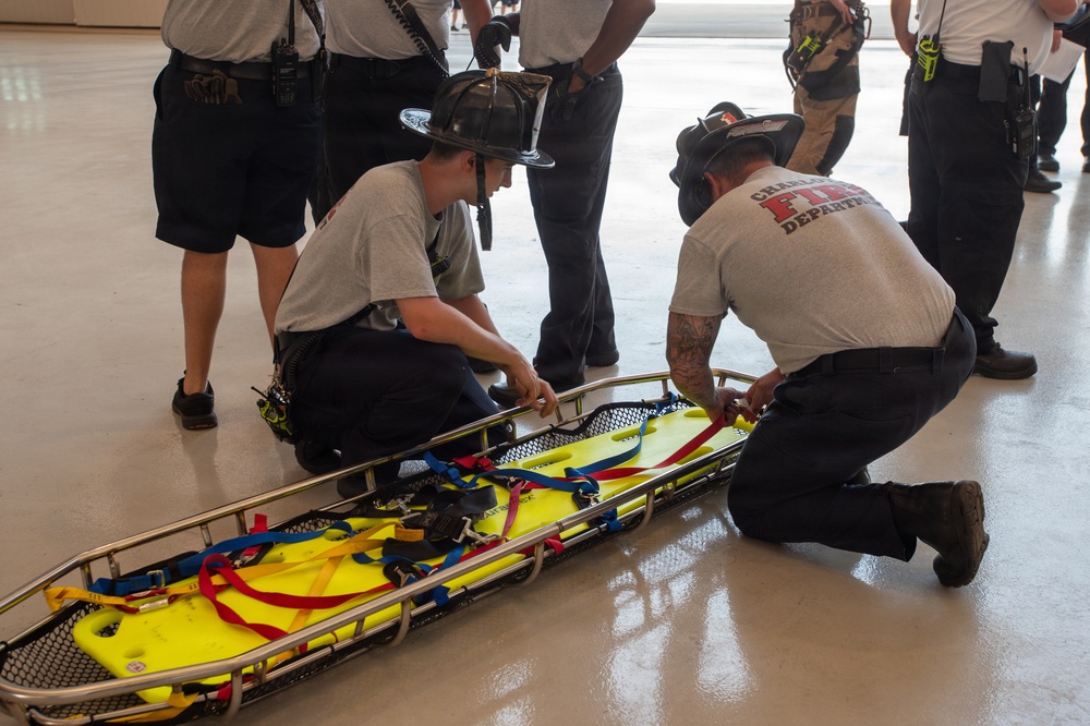 Two firefighters are kneeling on a polished concrete floor, working with a yellow Stokes basket stretcher equipped with blue and red restraint straps. The scene appears to be a training exercise, with multiple other uniformed personnel standing and observing in the background. One firefighter is wearing a helmet and the other has a shirt reading 'Charleston Fire Department'.