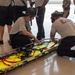 Two firefighters are kneeling on a polished concrete floor, working with a yellow Stokes basket stretcher equipped with blue and red restraint straps. The scene appears to be a training exercise, with multiple other uniformed personnel standing and observing in the background. One firefighter is wearing a helmet and the other has a shirt reading 'Charleston Fire Department'.
