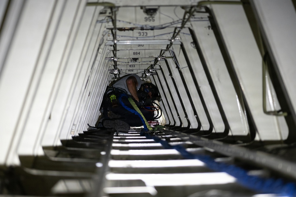 A technician or maintenance worker is crouched inside a narrow, elongated structural cavity, likely the interior of a large aircraft fuselage or cargo hold undergoing maintenance. The space features evenly spaced metal ribs/frames along both sides and ceiling, with numbered station markers visible on the walls. The worker appears to be performing hands-on technical work with tools and cables in a confined space.