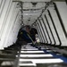 A technician or maintenance worker is crouched inside a narrow, elongated structural cavity, likely the interior of a large aircraft fuselage or cargo hold undergoing maintenance. The space features evenly spaced metal ribs/frames along both sides and ceiling, with numbered station markers visible on the walls. The worker appears to be performing hands-on technical work with tools and cables in a confined space.