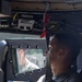 A U.S. Air Force service member in MultiCam/OCP camouflage uniform is seated inside a military HMMWV (Humvee), wearing a seatbelt and looking forward through the windshield. The interior shows military communications equipment mounted overhead and standard Humvee door panels with safety placards. The image captures the soldier from a rear-side profile perspective in what appears to be a training or operational environment.