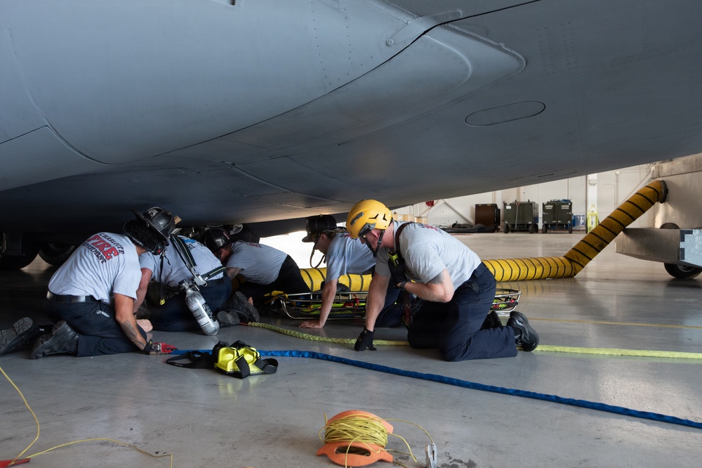 A team of firefighters or rescue personnel are conducting what appears to be an aircraft rescue training exercise beneath the fuselage of a large military aircraft inside a hangar. The crew is working to extract a simulated casualty using a rescue litter/basket stretcher in a confined space scenario. Multiple responders are kneeling and crouching under the aircraft, coordinating the extraction effort.