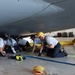 A team of firefighters or rescue personnel are conducting what appears to be an aircraft rescue training exercise beneath the fuselage of a large military aircraft inside a hangar. The crew is working to extract a simulated casualty using a rescue litter/basket stretcher in a confined space scenario. Multiple responders are kneeling and crouching under the aircraft, coordinating the extraction effort.