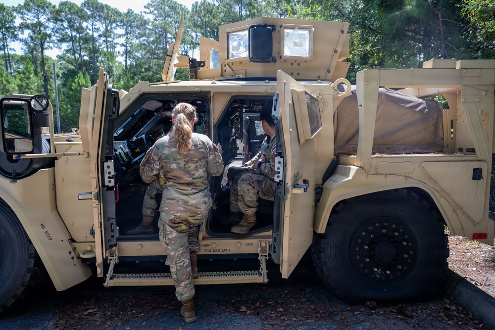 Two military personnel in MultiCam/OCP camouflage uniforms are interacting with a tan/desert-colored MRAP (Mine-Resistant Ambush Protected) vehicle. One soldier is standing outside facing the open door while a second soldier is seated inside the vehicle cabin. The setting appears to be a wooded training area or military installation.