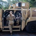 Two military personnel in MultiCam/OCP camouflage uniforms are interacting with a tan/desert-colored MRAP (Mine-Resistant Ambush Protected) vehicle. One soldier is standing outside facing the open door while a second soldier is seated inside the vehicle cabin. The setting appears to be a wooded training area or military installation.