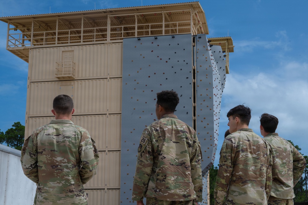 Four military personnel in OCP (Operational Camouflage Pattern) uniforms stand with their backs to the camera, observing a multi-story training tower facility. The structure features a climbing wall with holds on its face and appears to be a military obstacle course or training complex. The setting appears to be an outdoor military training installation surrounded by trees.
