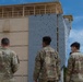 Four military personnel in OCP (Operational Camouflage Pattern) uniforms stand with their backs to the camera, observing a multi-story training tower facility. The structure features a climbing wall with holds on its face and appears to be a military obstacle course or training complex. The setting appears to be an outdoor military training installation surrounded by trees.