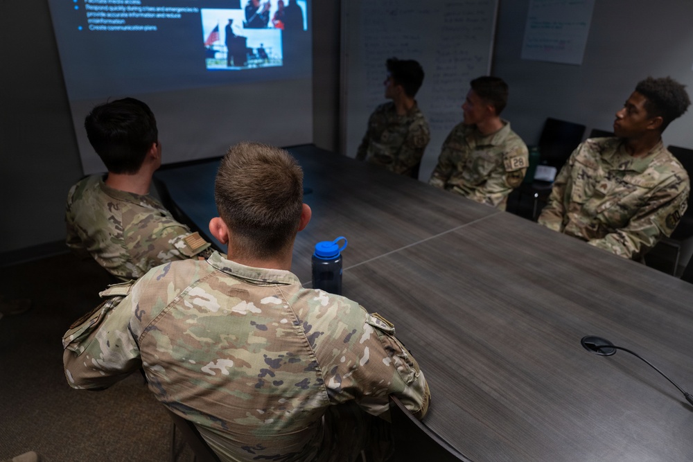 A group of military personnel in OCP (Operational Camouflage Pattern) uniforms are seated around a conference table in a briefing room. They are attending a presentation displayed on a projected screen at the front of the room. The setting appears to be a formal military training or briefing session.