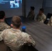 A group of military personnel in OCP (Operational Camouflage Pattern) uniforms are seated around a conference table in a briefing room. They are attending a presentation displayed on a projected screen at the front of the room. The setting appears to be a formal military training or briefing session.