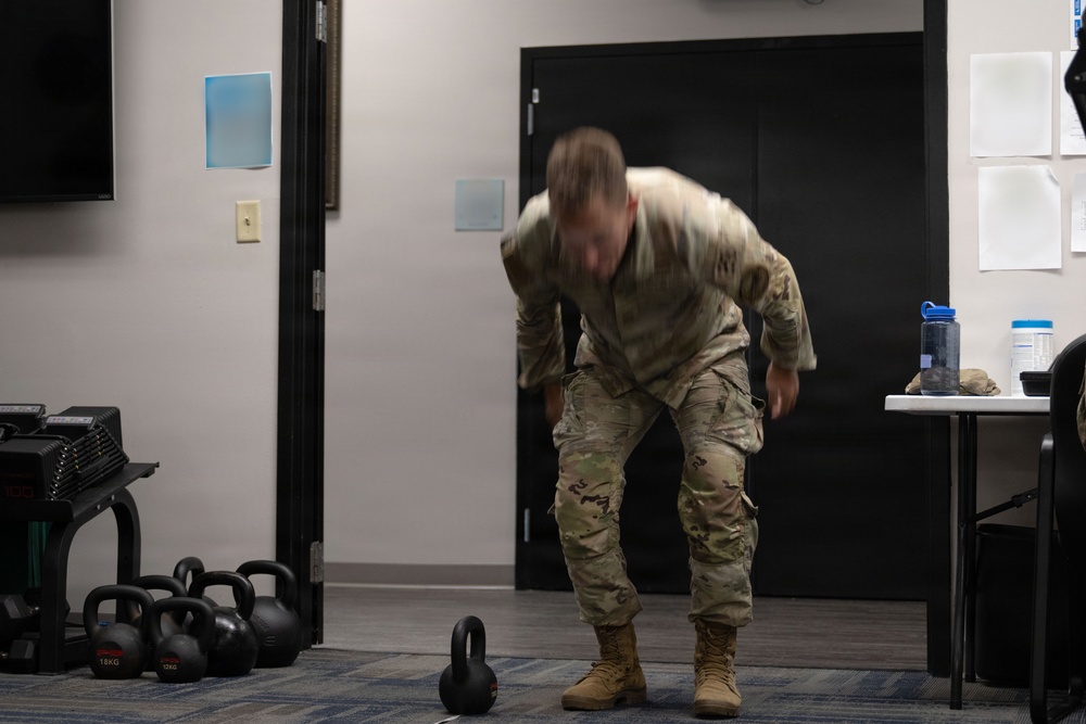 A person wearing military camouflage uniform and tan combat boots is performing a kettlebell exercise in what appears to be an improvised fitness area inside a building. The individual is bent forward in motion, likely executing a kettlebell swing or deadlift movement, with motion blur indicating active movement. The room contains multiple kettlebells of varying weights, a dumbbell rack, and a table with water bottles.