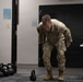 A person wearing military camouflage uniform and tan combat boots is performing a kettlebell exercise in what appears to be an improvised fitness area inside a building. The individual is bent forward in motion, likely executing a kettlebell swing or deadlift movement, with motion blur indicating active movement. The room contains multiple kettlebells of varying weights, a dumbbell rack, and a table with water bottles.