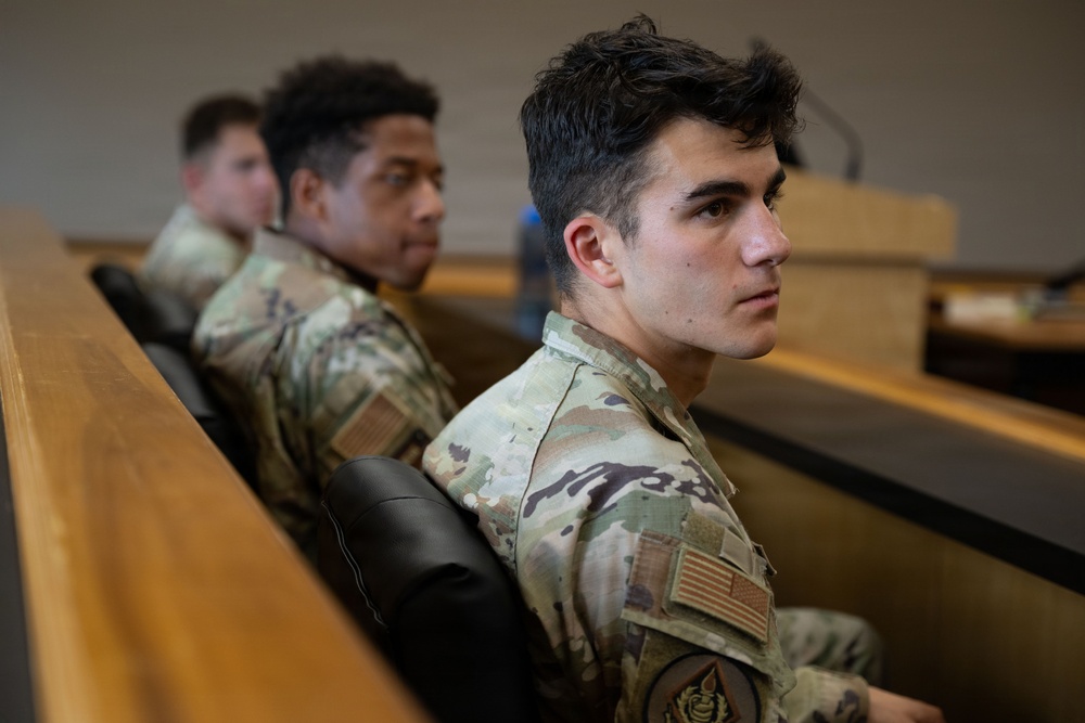 Three individuals wearing U.S. Army OCP (Operational Camouflage Pattern) uniforms are seated in what appears to be a classroom or briefing room with tiered seating. The foreground subject is in sharp focus, looking attentively to the right, while the two individuals behind are progressively out of focus. An American flag patch and a unit patch are visible on the foreground subject's uniform.
