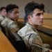 Three individuals wearing U.S. Army OCP (Operational Camouflage Pattern) uniforms are seated in what appears to be a classroom or briefing room with tiered seating. The foreground subject is in sharp focus, looking attentively to the right, while the two individuals behind are progressively out of focus. An American flag patch and a unit patch are visible on the foreground subject's uniform.