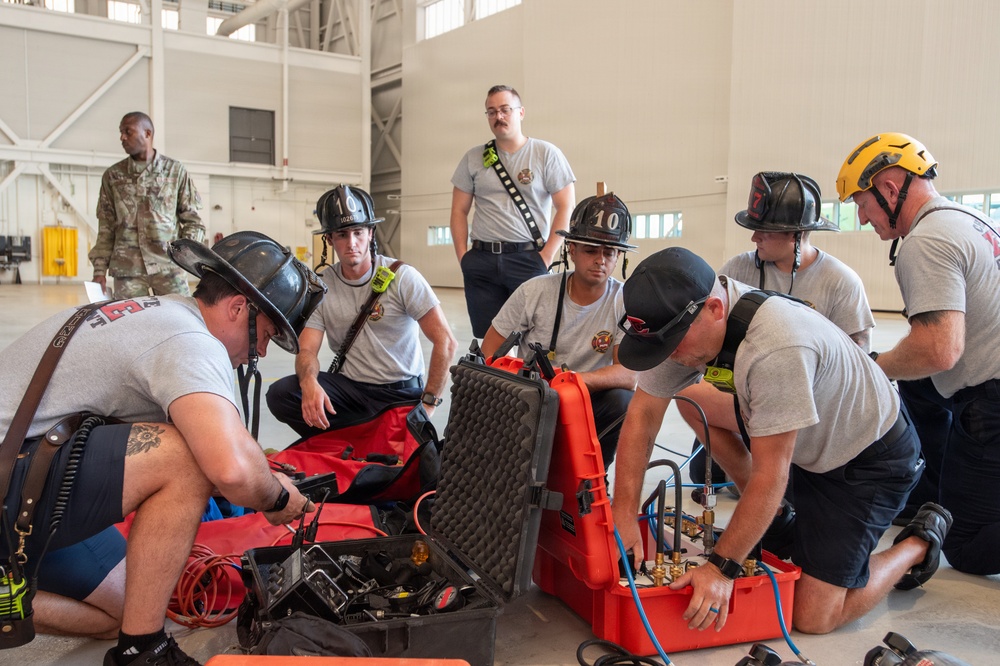 A group of firefighters in gray t-shirts and traditional black helmets are gathered on the floor of what appears to be a large hangar or training facility, examining and working with specialized equipment cases. The red hard-shell cases contain what appear to be pneumatic or hydraulic tools with gauges and fittings. A military personnel member in camouflage uniform is visible in the background, suggesting this may be a joint training exercise at a military installation.