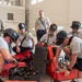 A group of firefighters in gray t-shirts and traditional black helmets are gathered on the floor of what appears to be a large hangar or training facility, examining and working with specialized equipment cases. The red hard-shell cases contain what appear to be pneumatic or hydraulic tools with gauges and fittings. A military personnel member in camouflage uniform is visible in the background, suggesting this may be a joint training exercise at a military installation.