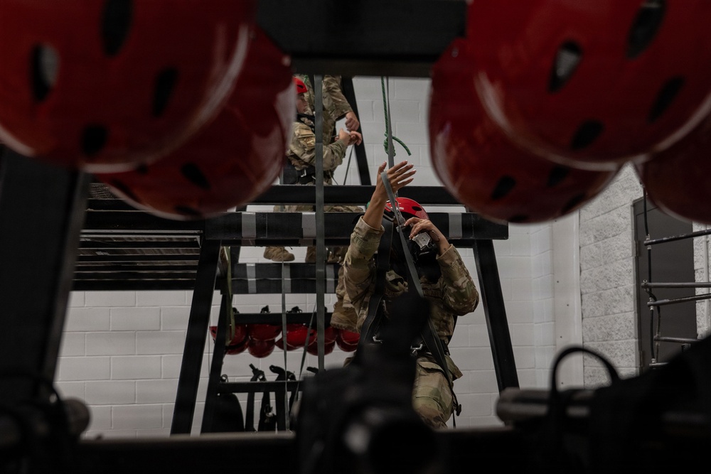 Military personnel in camouflage uniforms and red helmets are engaged in an indoor rope or rappelling training exercise. The image is taken from a low angle between two large red helmets in the foreground, showing at least two individuals working with ropes and rigging equipment on a metal training structure. The setting appears to be an indoor training facility with white cinder block walls.