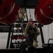 Military personnel in camouflage uniforms and red helmets are engaged in an indoor rope or rappelling training exercise. The image is taken from a low angle between two large red helmets in the foreground, showing at least two individuals working with ropes and rigging equipment on a metal training structure. The setting appears to be an indoor training facility with white cinder block walls.