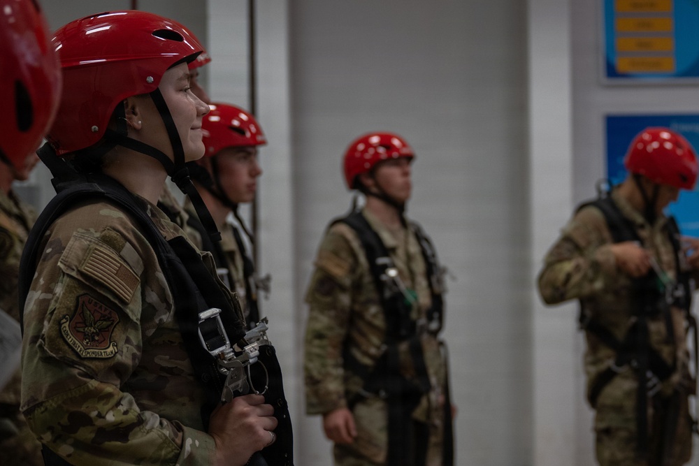 A group of military personnel wearing OCP (Operational Camouflage Pattern) uniforms are equipped with red helmets and rappelling/climbing harnesses, standing in a line inside what appears to be a training facility. The foreground individual displays a US flag patch and what appears to be a unit patch on their left shoulder. They appear to be preparing for or awaiting a rappelling or aerial training exercise.