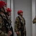 A group of military personnel wearing OCP (Operational Camouflage Pattern) uniforms are equipped with red helmets and rappelling/climbing harnesses, standing in a line inside what appears to be a training facility. The foreground individual displays a US flag patch and what appears to be a unit patch on their left shoulder. They appear to be preparing for or awaiting a rappelling or aerial training exercise.