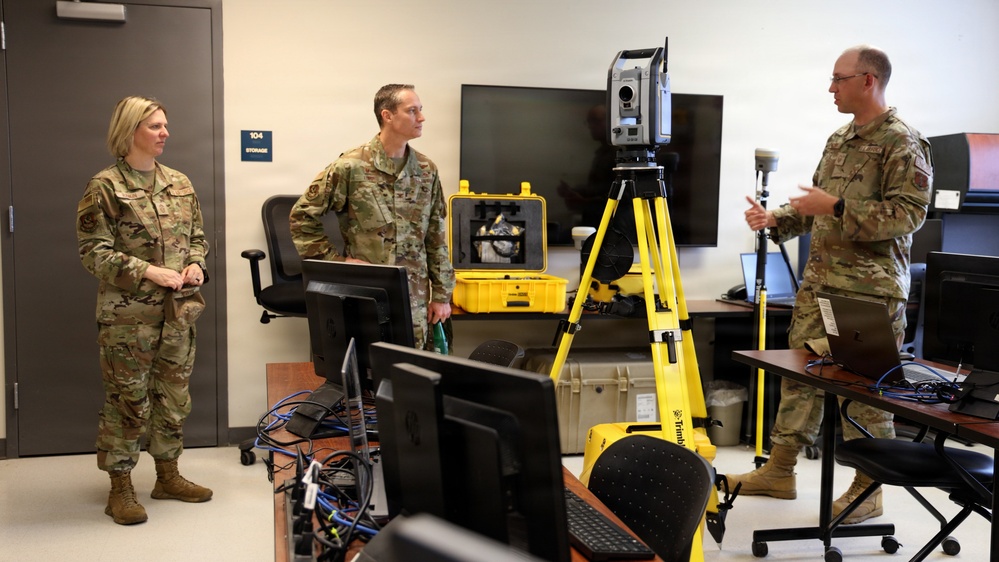 Three military personnel in OCP (Operational Camouflage Pattern) uniforms are gathered in what appears to be a technical laboratory or training room. One individual on the right appears to be briefing or demonstrating equipment to the other two. A Trimble surveying total station on a yellow tripod is prominently displayed in the foreground.